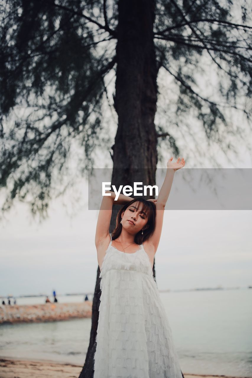 Portrait of young woman standing with arm raised against tree