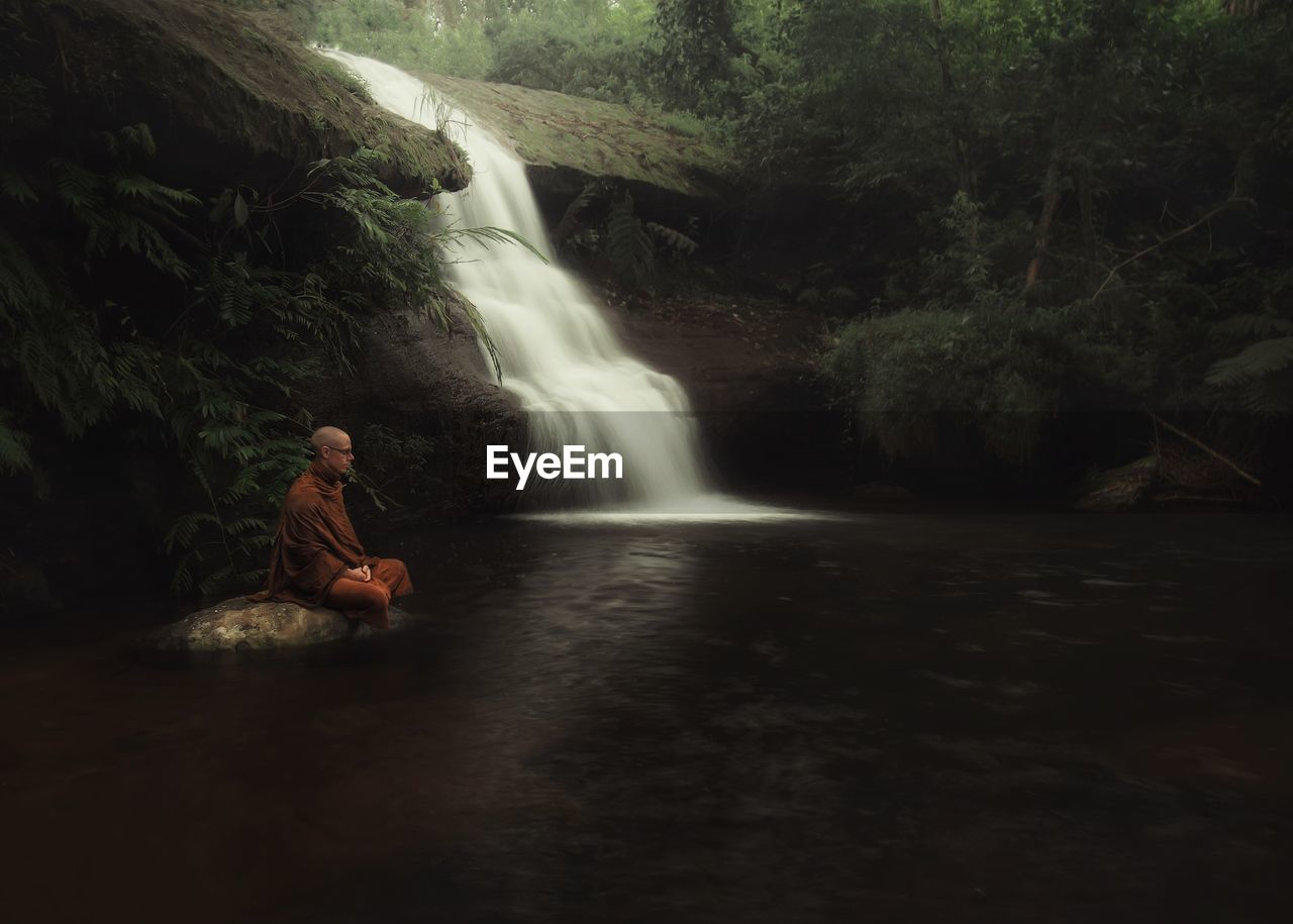 Side view of monk meditating against waterfall in forest
