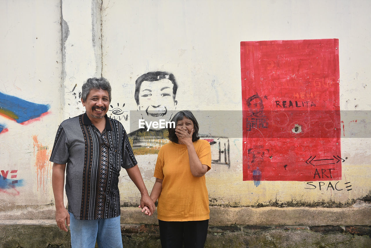 Portrait of mature couple holding hands while standing against graffiti wall