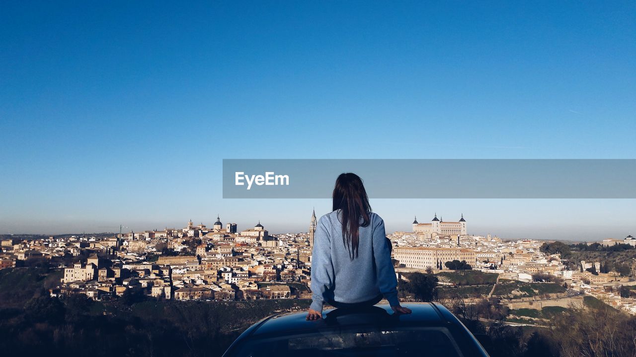 Rear view of woman looking at cityscape while sitting on car roof against clear blue sky