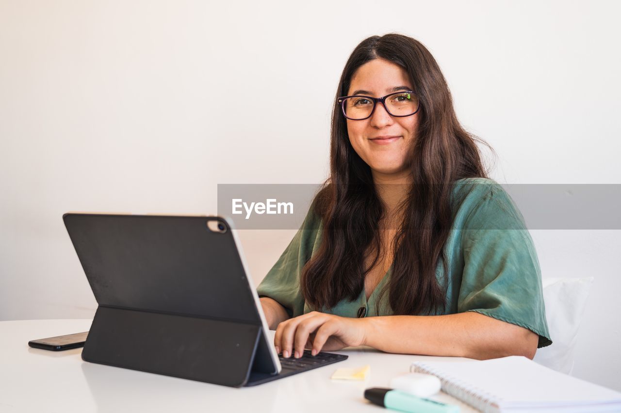 Young spanish brunette wearing glasses teleworking with her hybrid tablet laptop adapting her business to the new normal after the covid 19 global pandemic. she looks at camera and smiles
