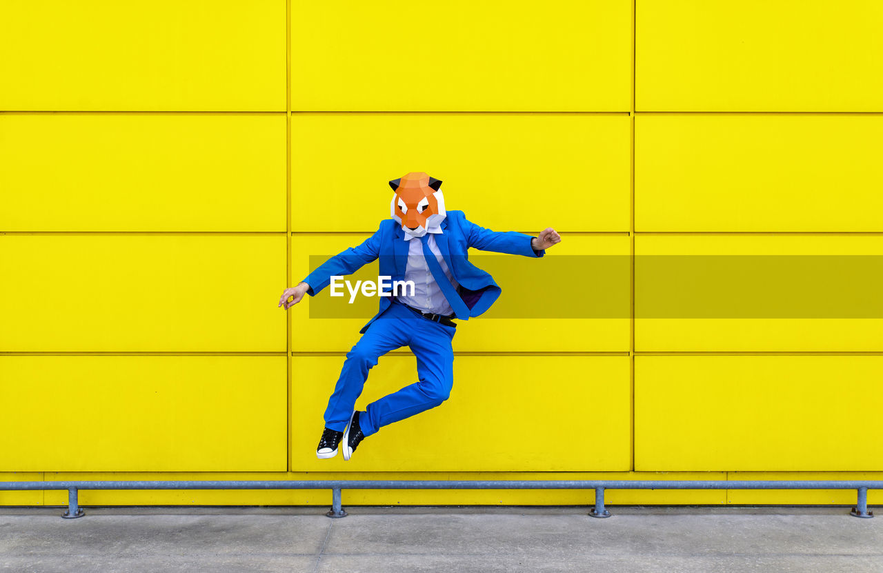 Man wearing vibrant blue suit and tiger mask jumping against yellow wall