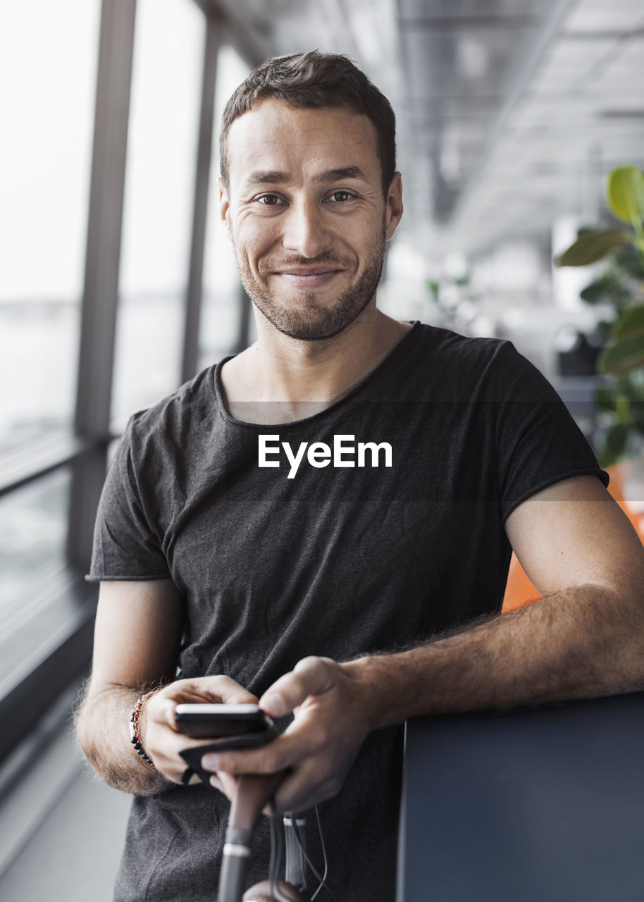 Portrait of smiling businessman holding mobile phone while leaning on cubicle in office