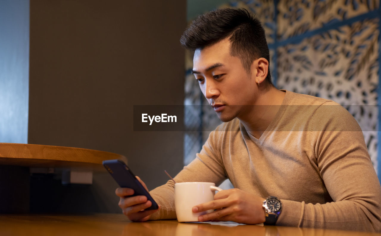 Thoughtful asian male relaxing in armchair in cozy coffee shop and enjoying hot beverage while browsing smartphone