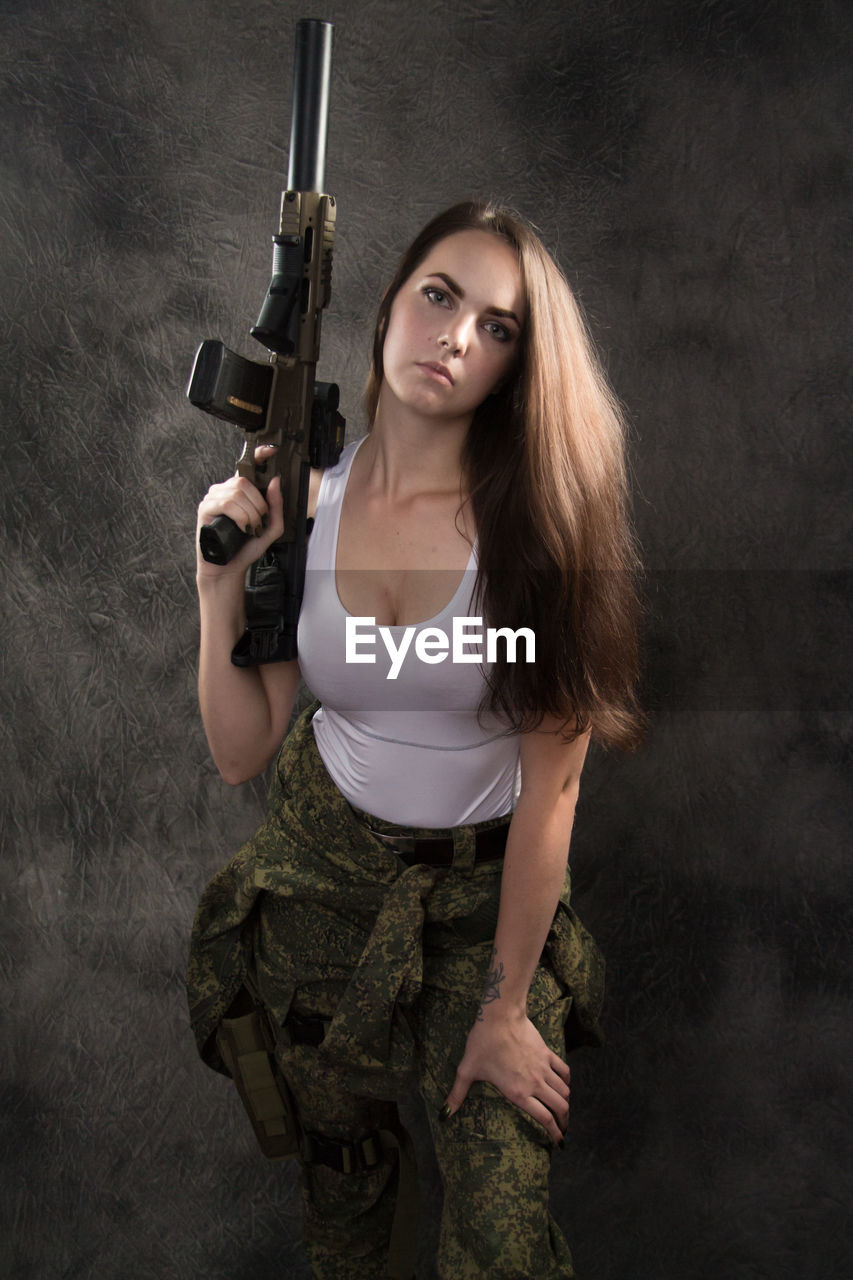 Portrait of beautiful young woman holding gun while standing against wall