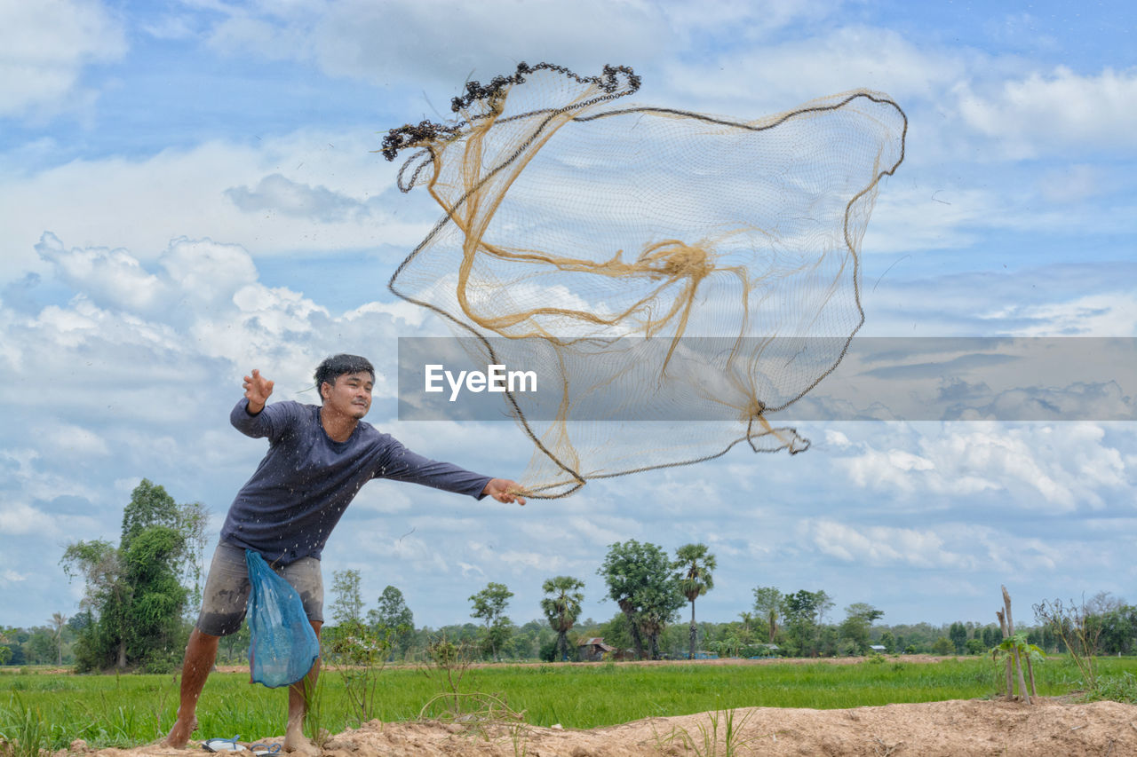 Full length of man throwing fishing net while standing on land against sky