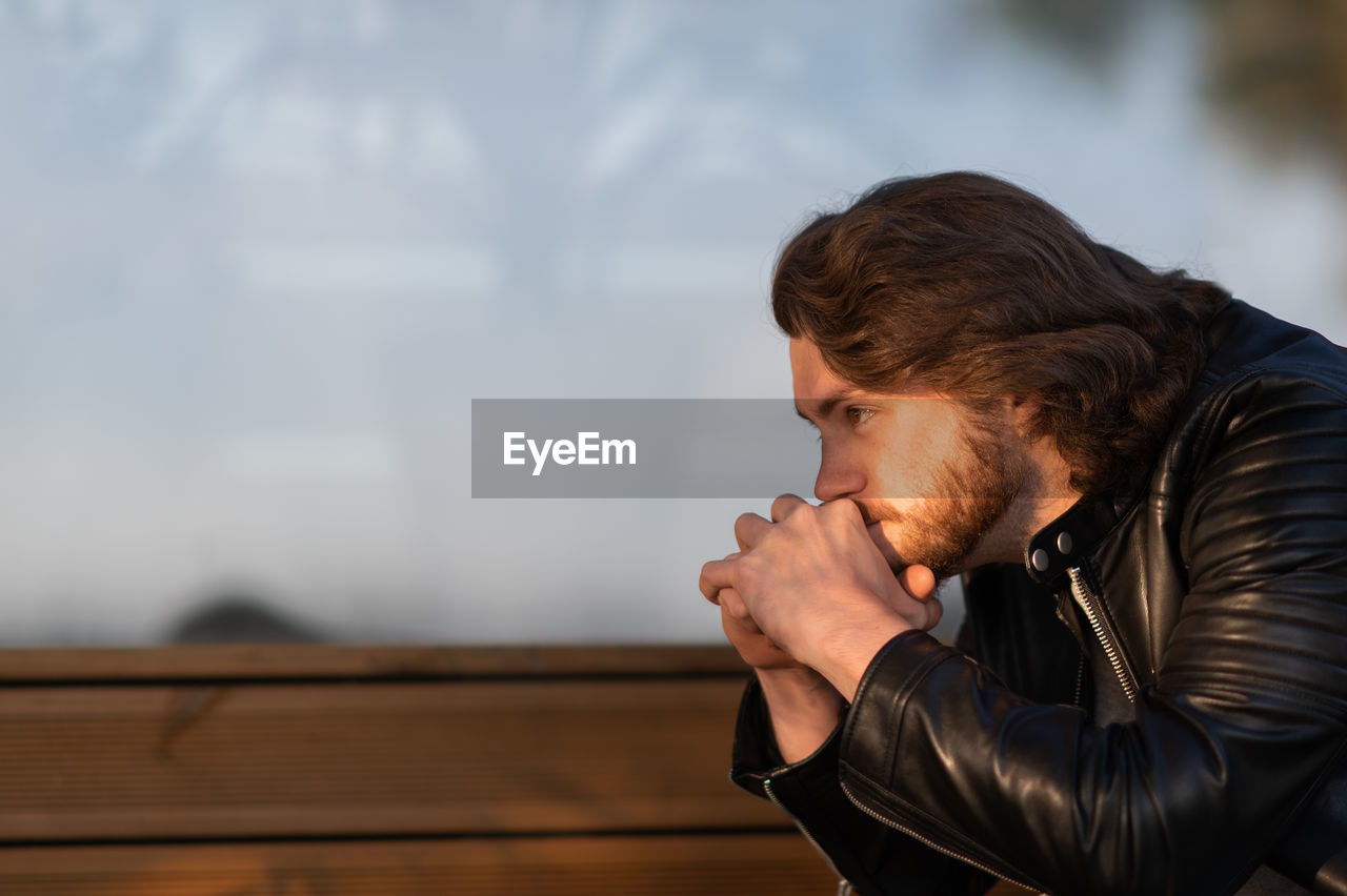 Pensive serious young bearded man wear black leather jacket sitting and thinking outdoors