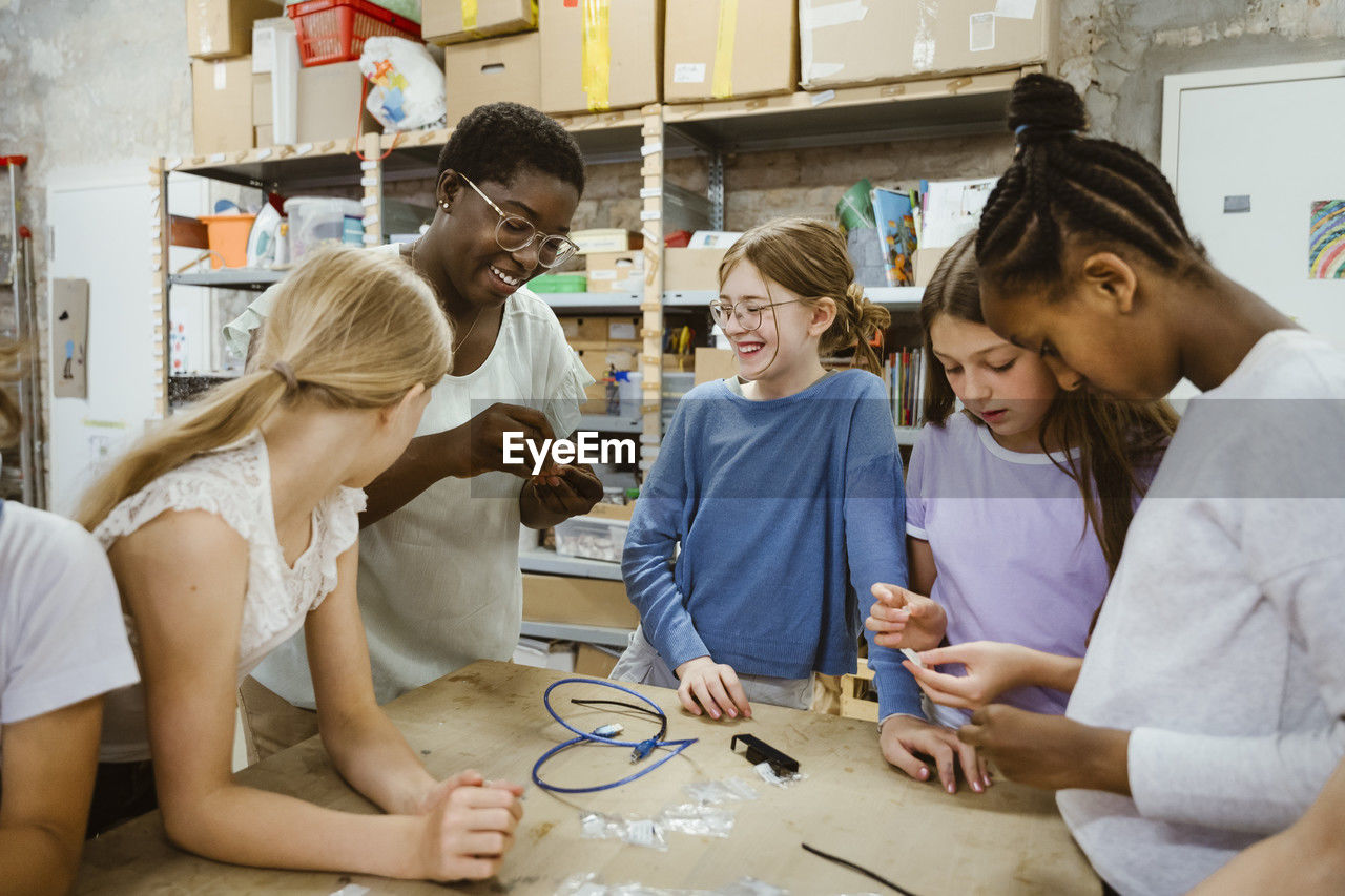 Female students with teacher holding electric component near table in workshop at school