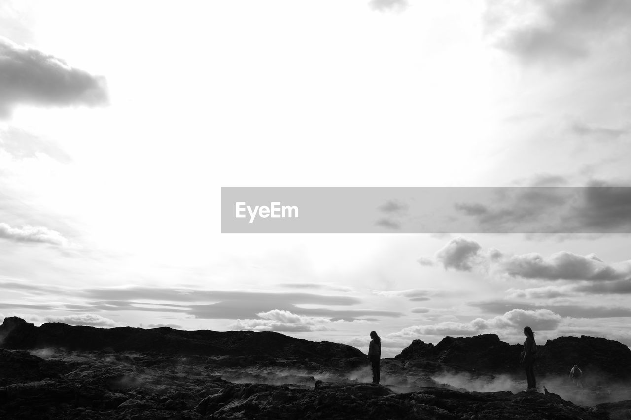 Friends standing on volcanic island against cloudy sky during sunset