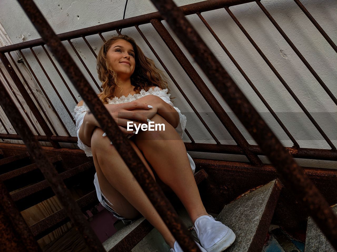 YOUNG WOMAN LOOKING AWAY WHILE SITTING ON RAILING AGAINST WINDOW