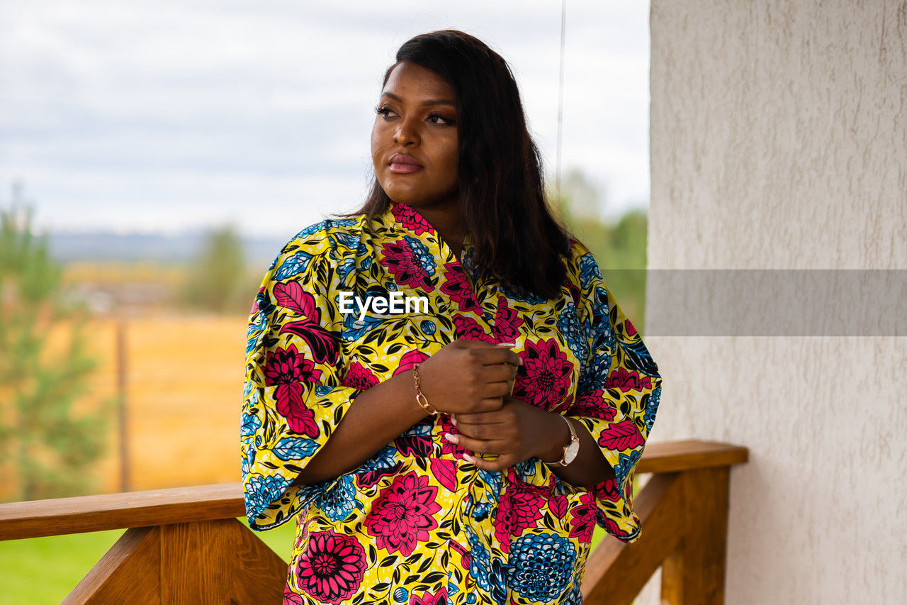 one person, adult, women, yellow, spring, standing, young adult, portrait, dress, architecture, photo shoot, hairstyle, lifestyles, waist up, clothing, floral pattern, looking, nature, three quarter length, pattern, female, emotion, outdoors, front view, contemplation, serious, day, fashion, smiling, looking at camera, person, relaxation, casual clothing, copy space, multi colored, traditional clothing, leisure activity, long hair, focus on foreground