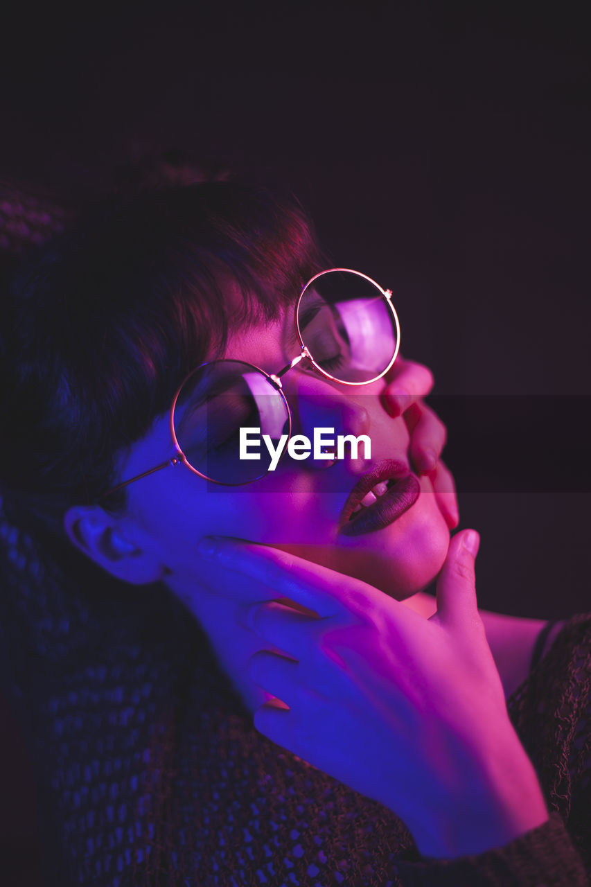 Tranquil female in stylish glasses touching face while standing with closed eyes on dark background in pink and blue neon illumination