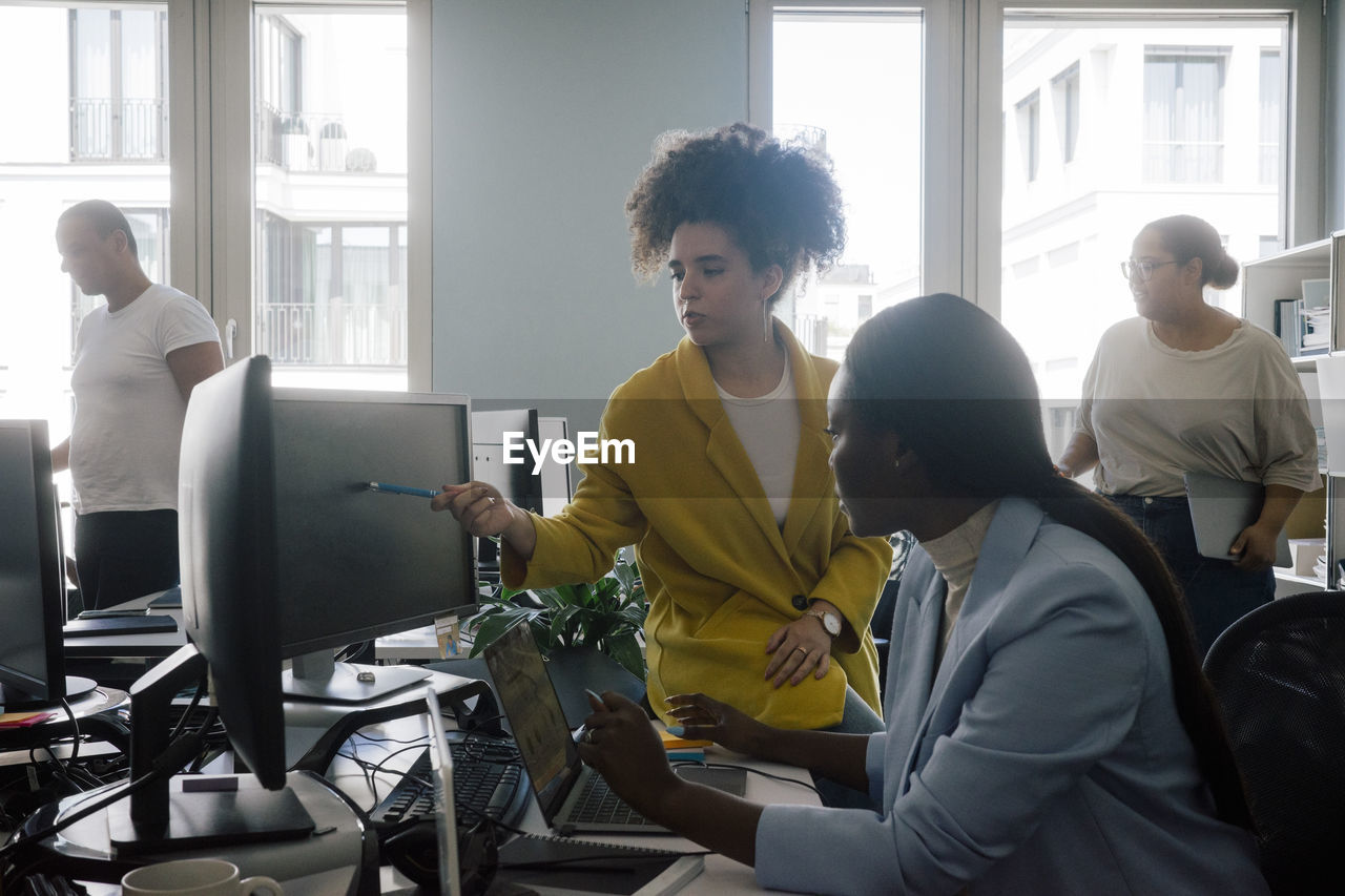 Businesswoman explaining coding to female colleague sitting at desk in office