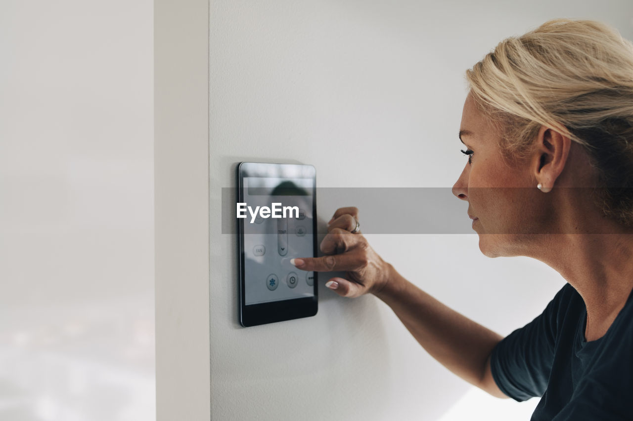 Blond woman adjusting thermostat using digital tablet mounted on white wall at home