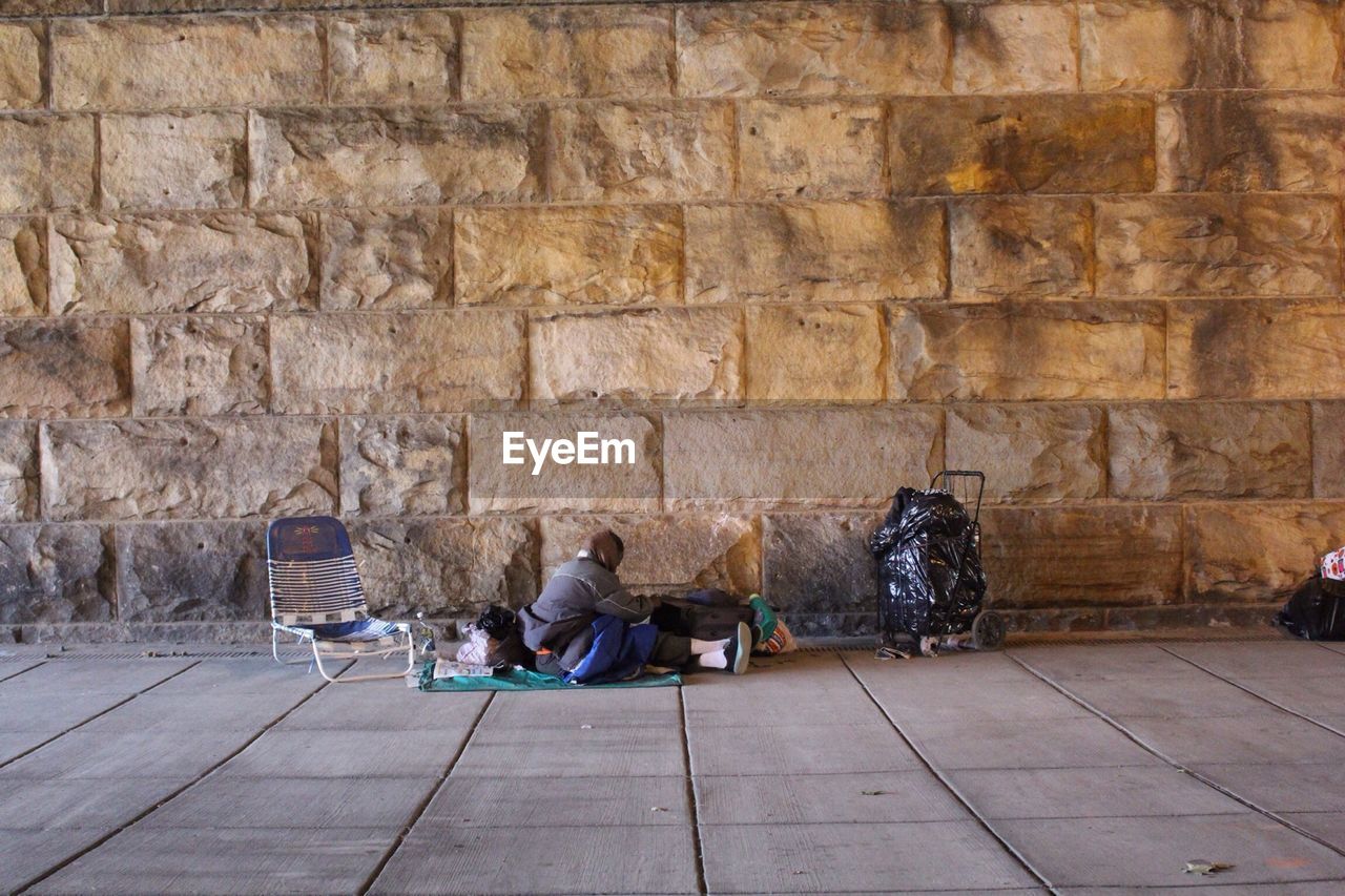 Homeless person sitting on sidewalk against stone wall