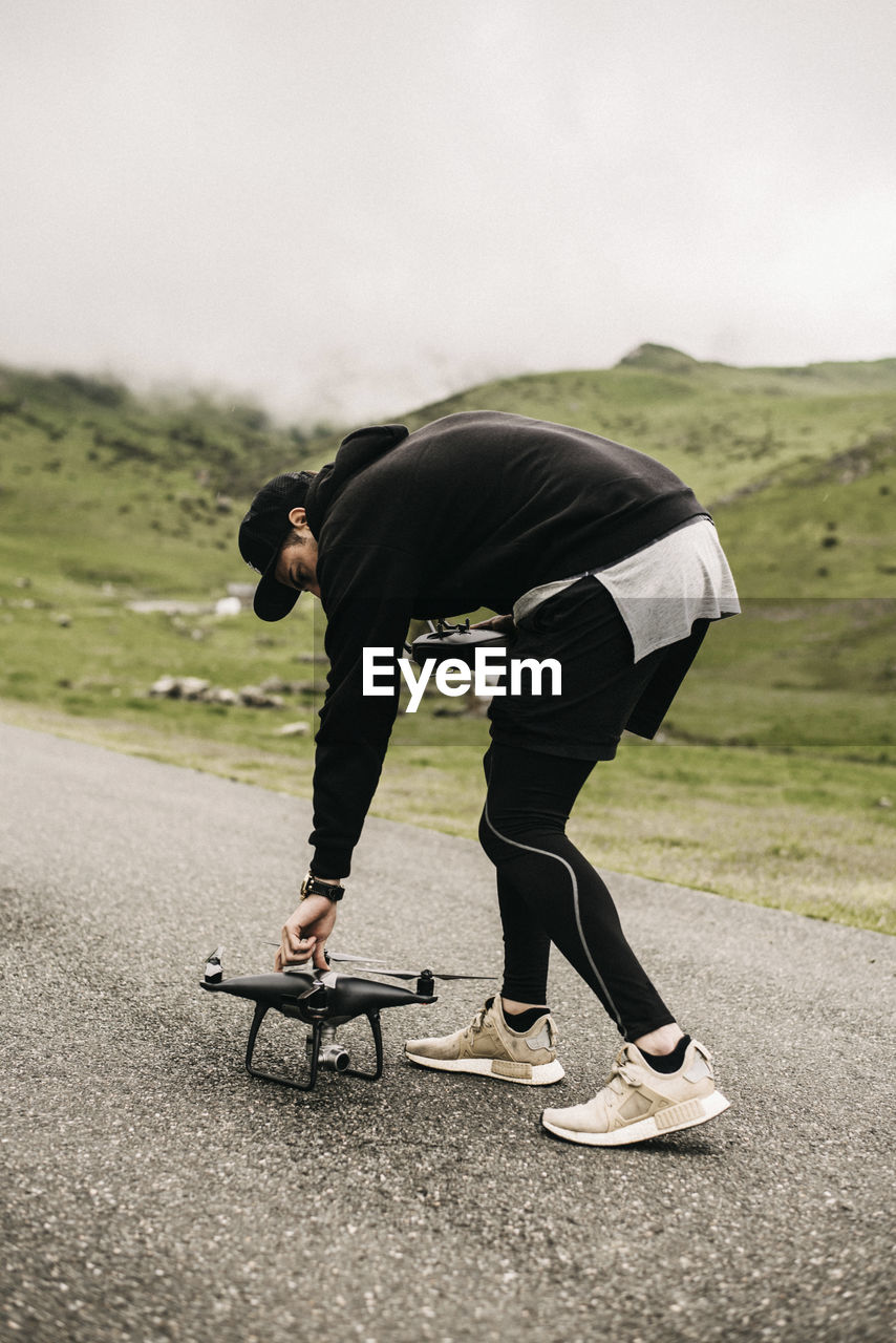 Side view of man holding quadcopter while standing on road against sky