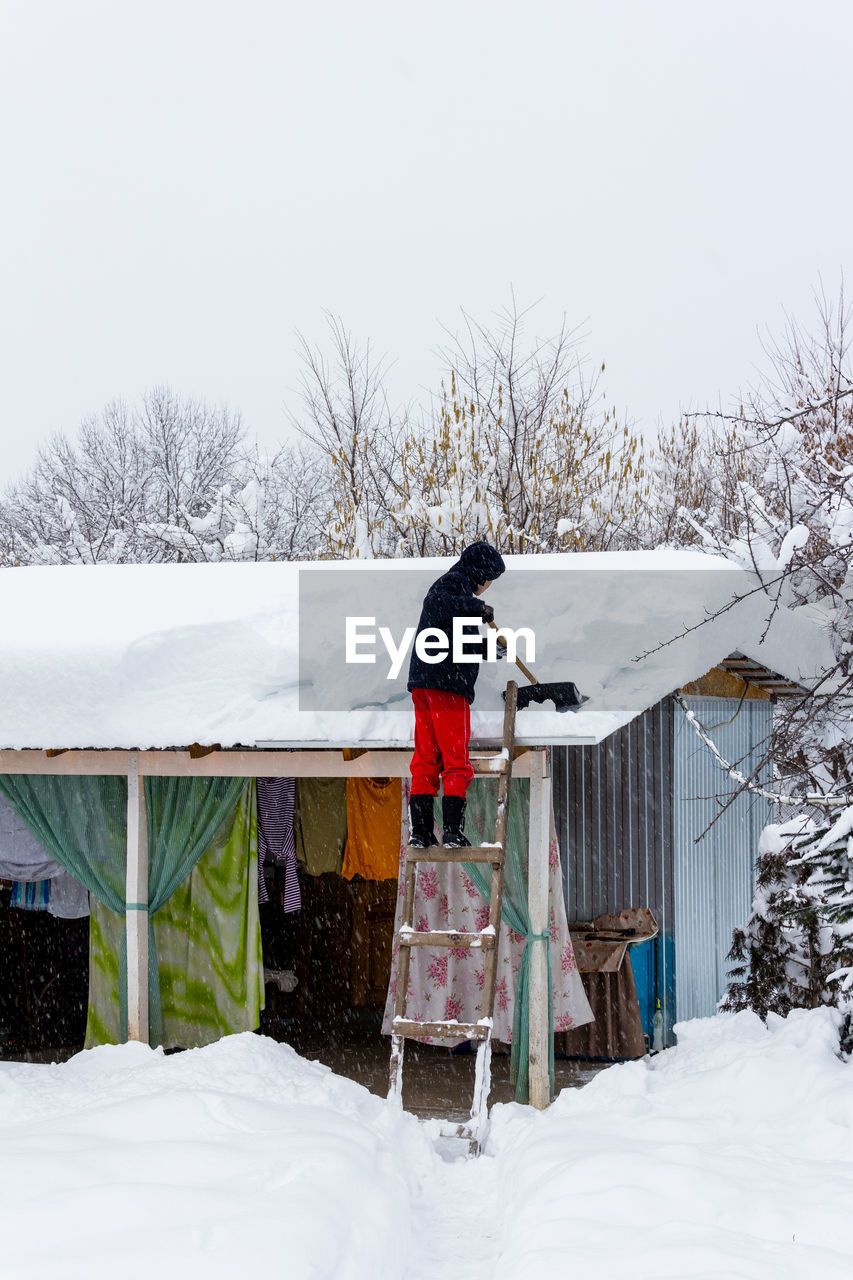 A guy shovels snow off a roof on a winter day in a snowfall