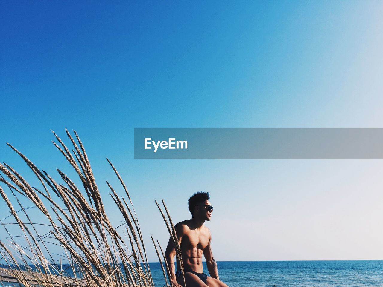 Shirtless young man sitting at beach against clear sky during sunny day
