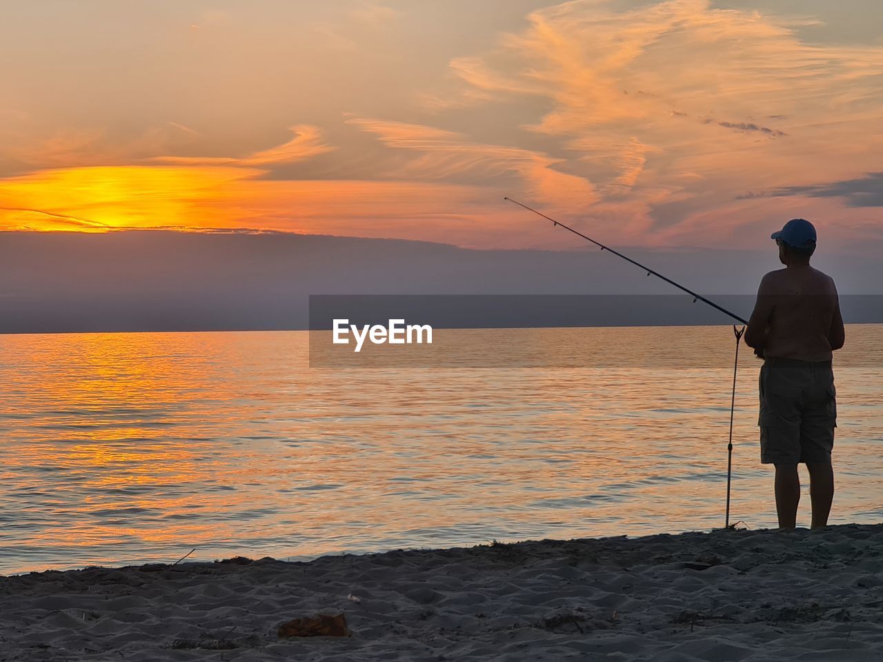 REAR VIEW OF MAN FISHING AT BEACH AGAINST SKY