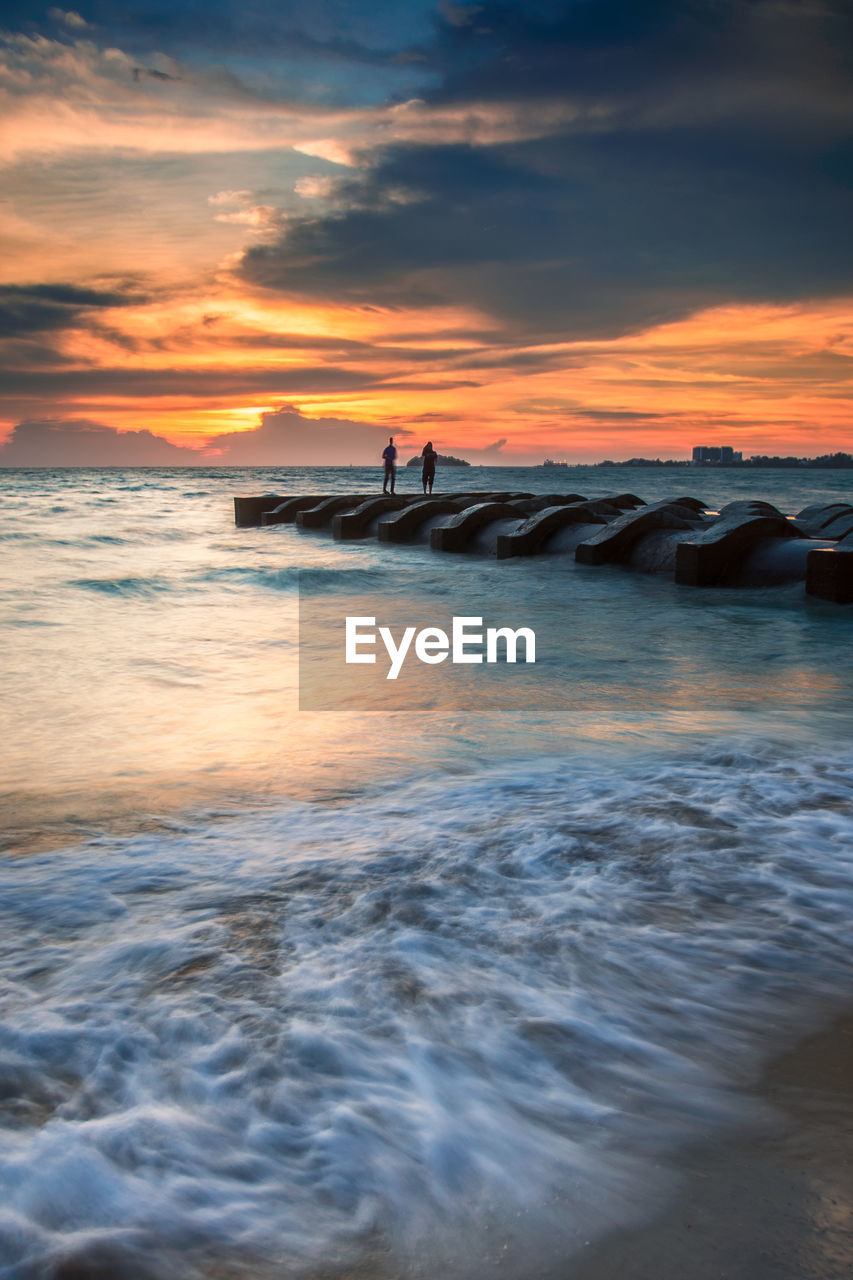 Silhouette of people standing on drainage pip in the sea at sunset