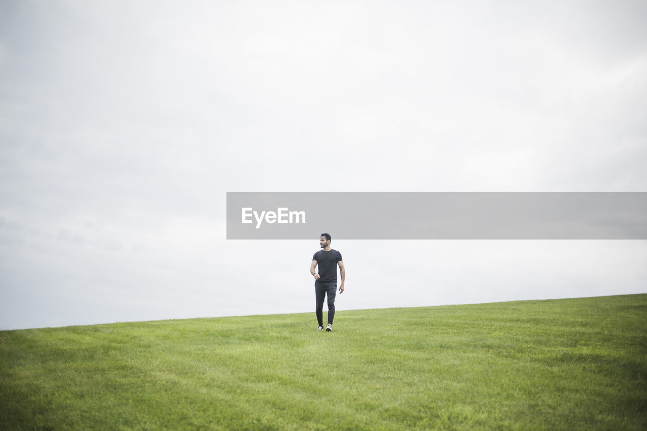 Mid distance view of young man walking on field against cloudy sky