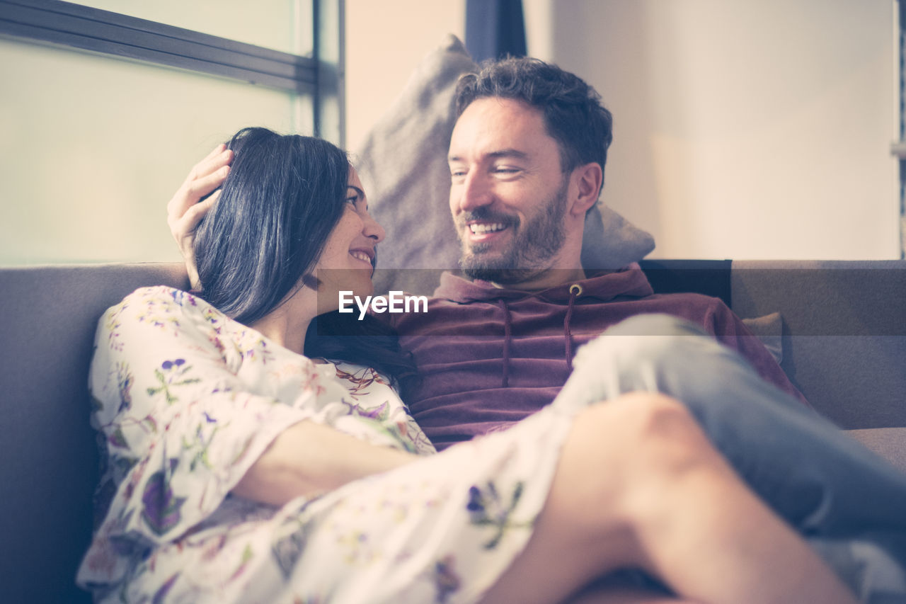 Smiling couple looking each other face to face while sitting on sofa at home
