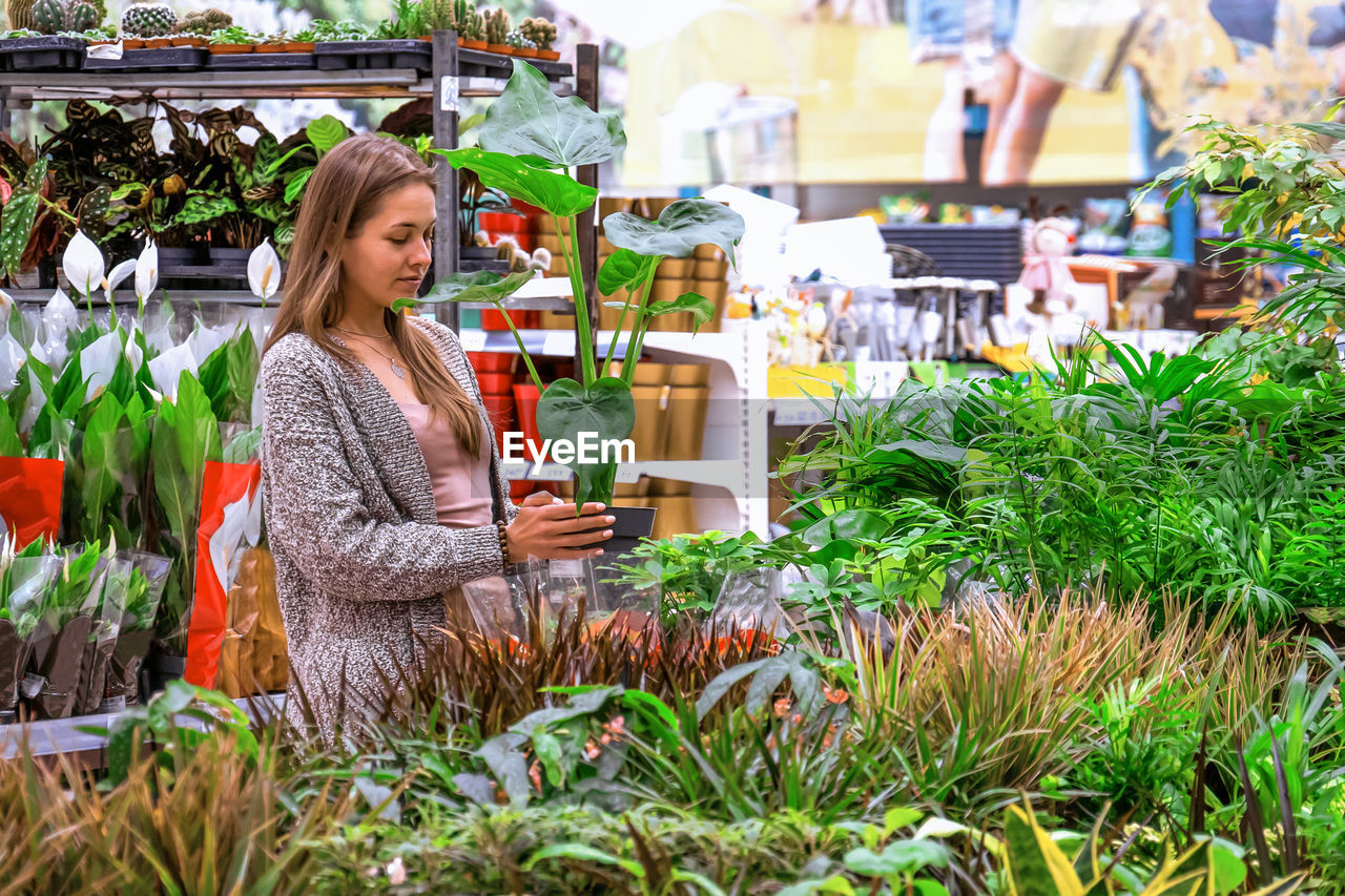 Young woman holding a pot with monstera in the middle of a flower store.