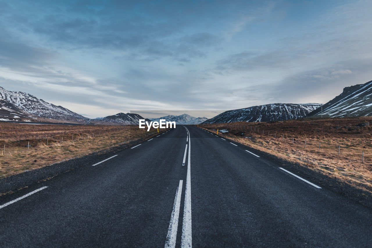 Empty road leading towards mountains against cloudy sky during winter