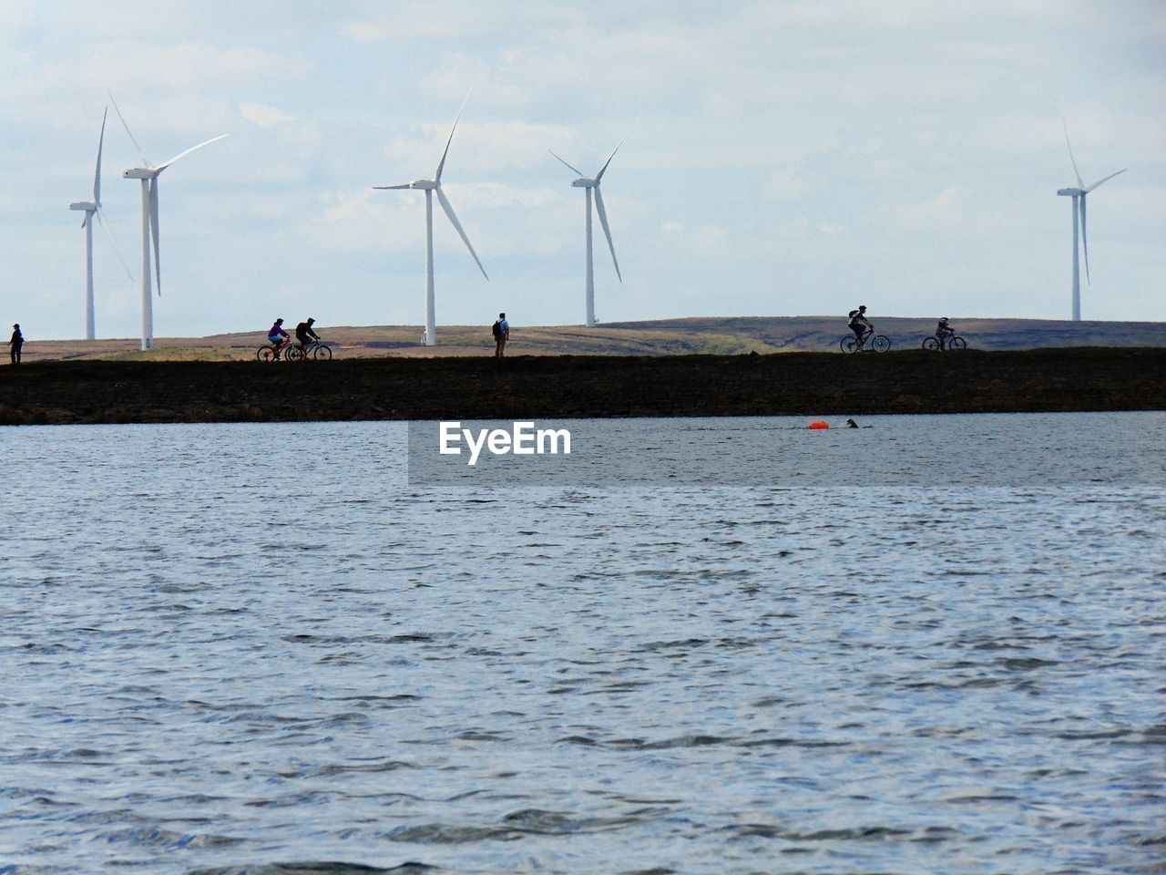 People riding bicycle on riverbank by windmills against sky