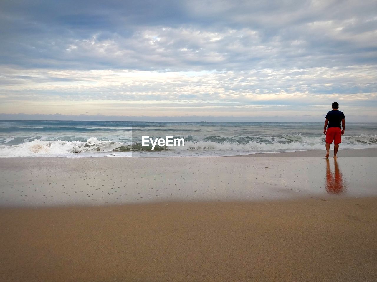 Rear view of man standing at beach against cloudy sky