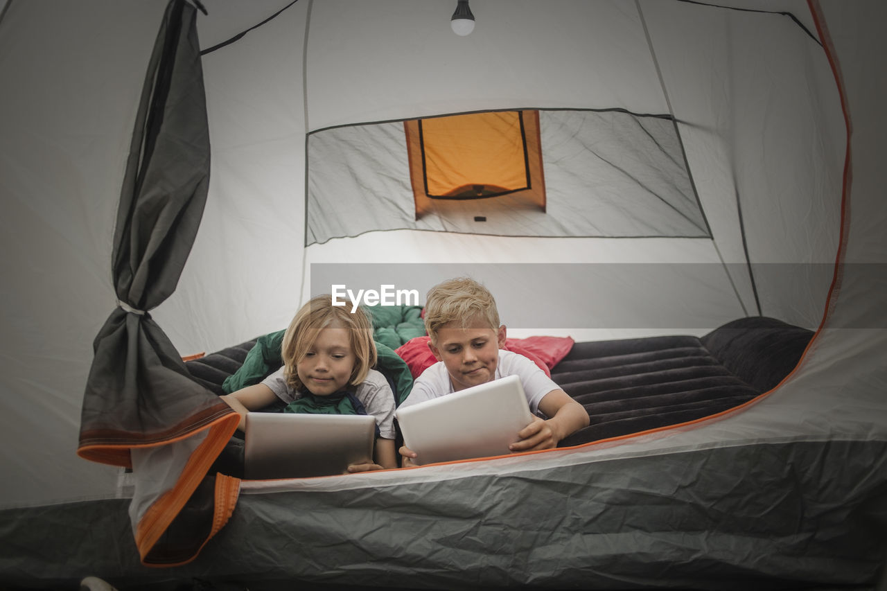 Siblings using digital tablet while lying together in tent at camping site