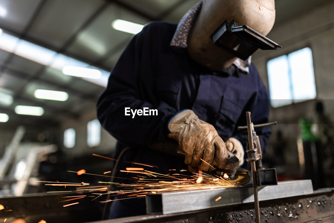 Unrecognizable male employee in protective gloves and helmet using welding machine while working in dark workshop