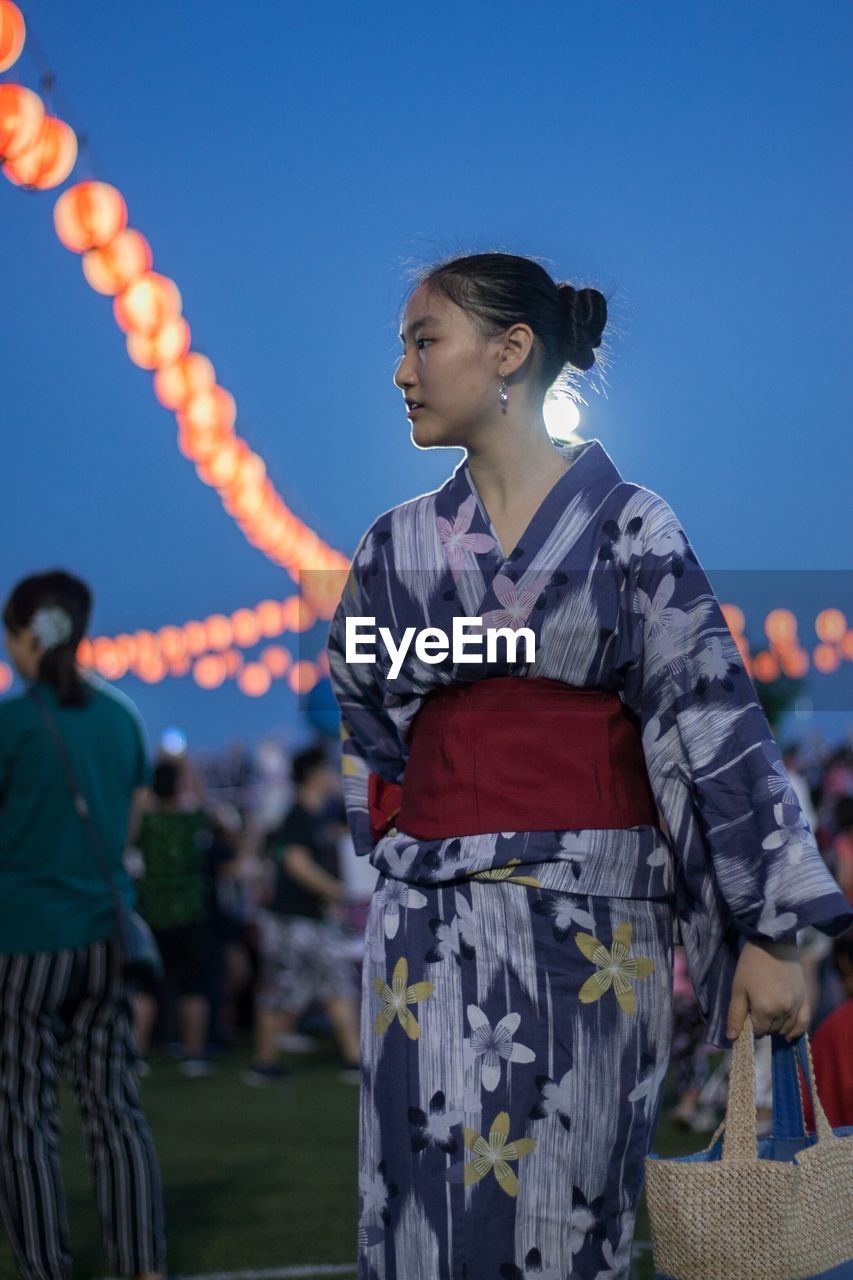 Woman wearing traditional clothing while standing against illuminated lanterns and sky at night