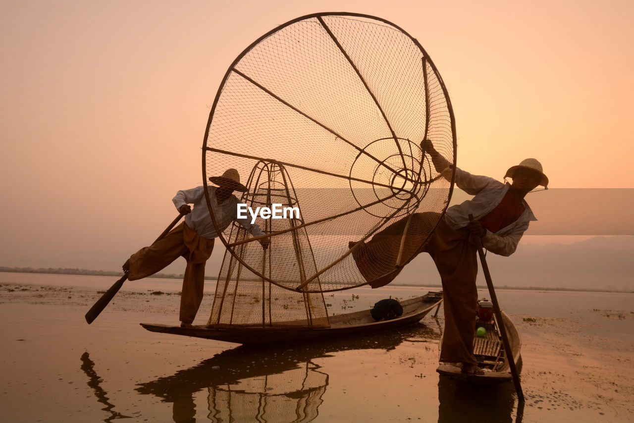 Fishermen fishing in lake against sky during sunset