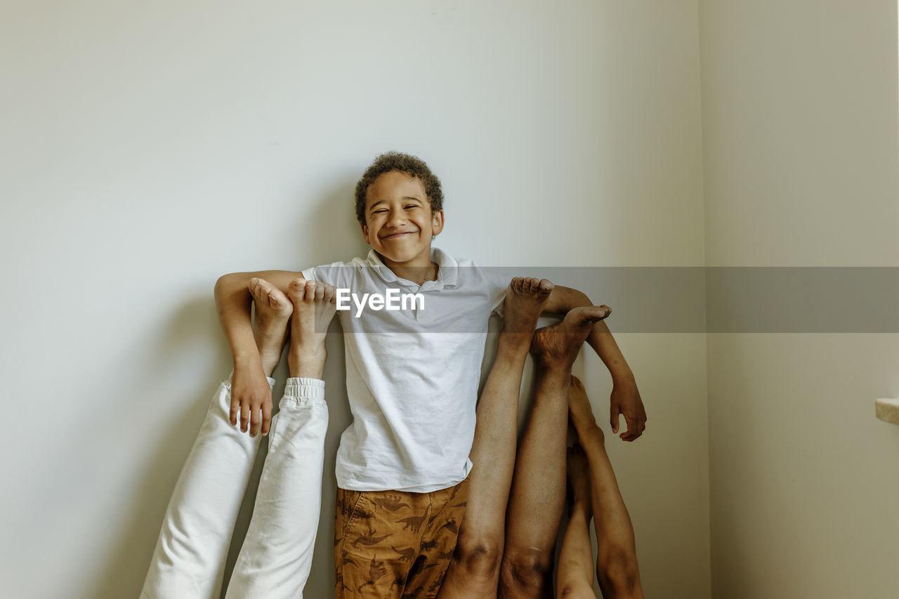 Happy boy with hands on parent's and sister's legs in front of wall