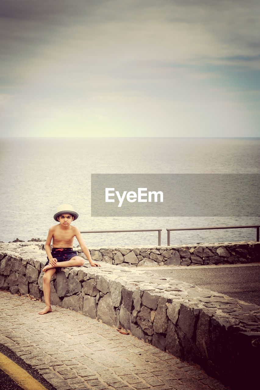 Shirtless boy looking away while sitting on retaining stone wall against sea
