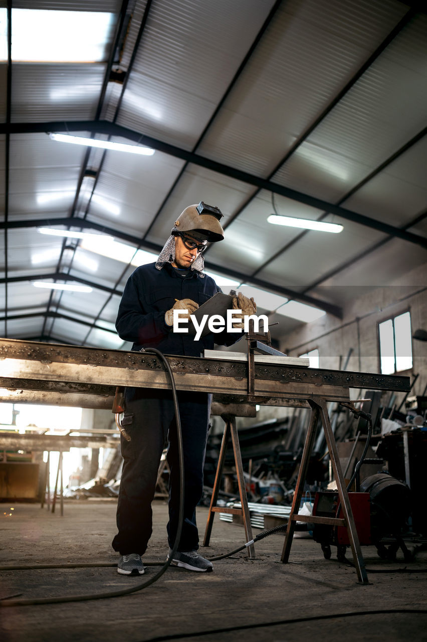 Serious male worker in hardhat and overall taking notes in notebook with pen on table near metal constructions in light garage