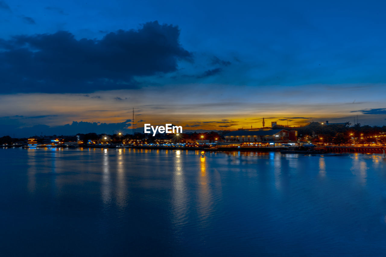 Illuminated city by sea against sky at sunset