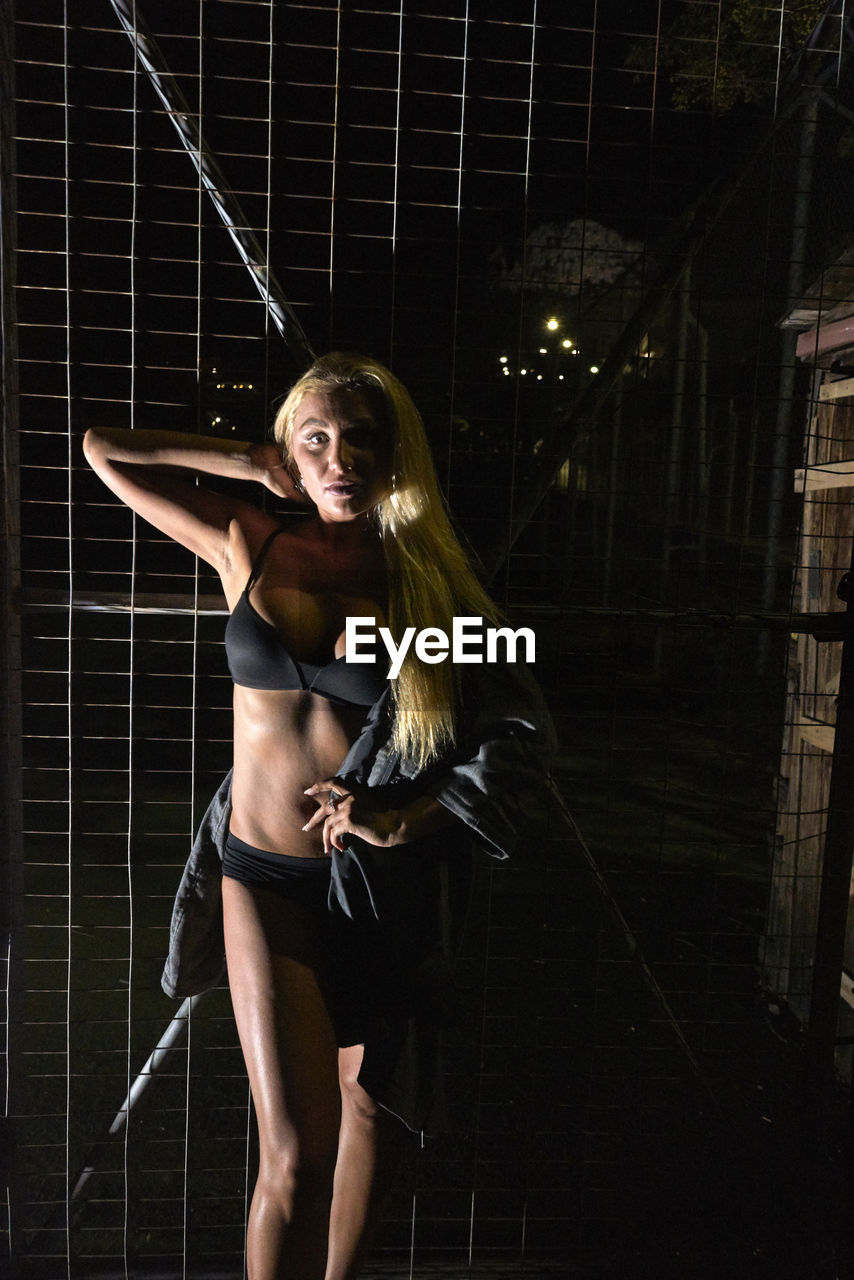 Seductive woman wearing black bikini while standing against metal grate in darkroom