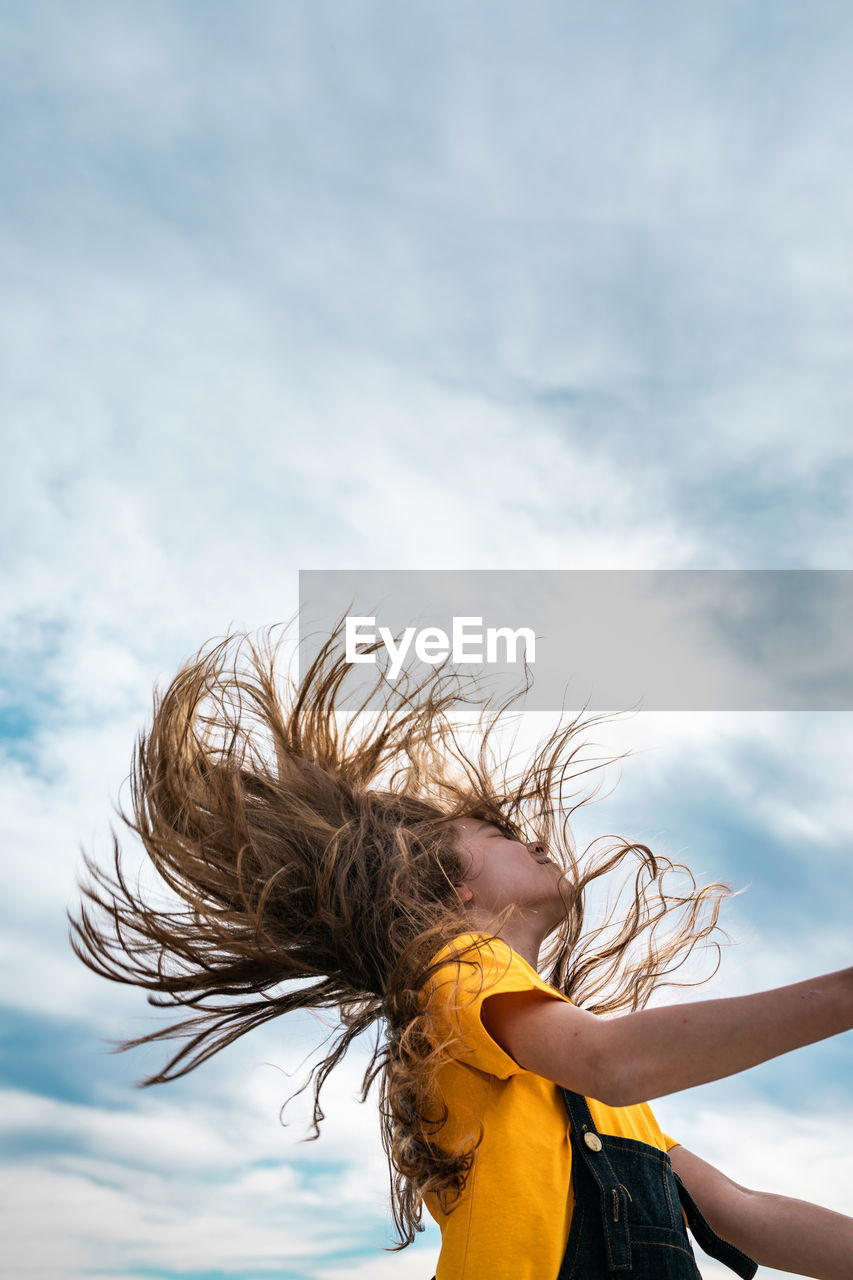 Side view from below of peaceful teenager throwing long hair on background of cloudy sky in summer