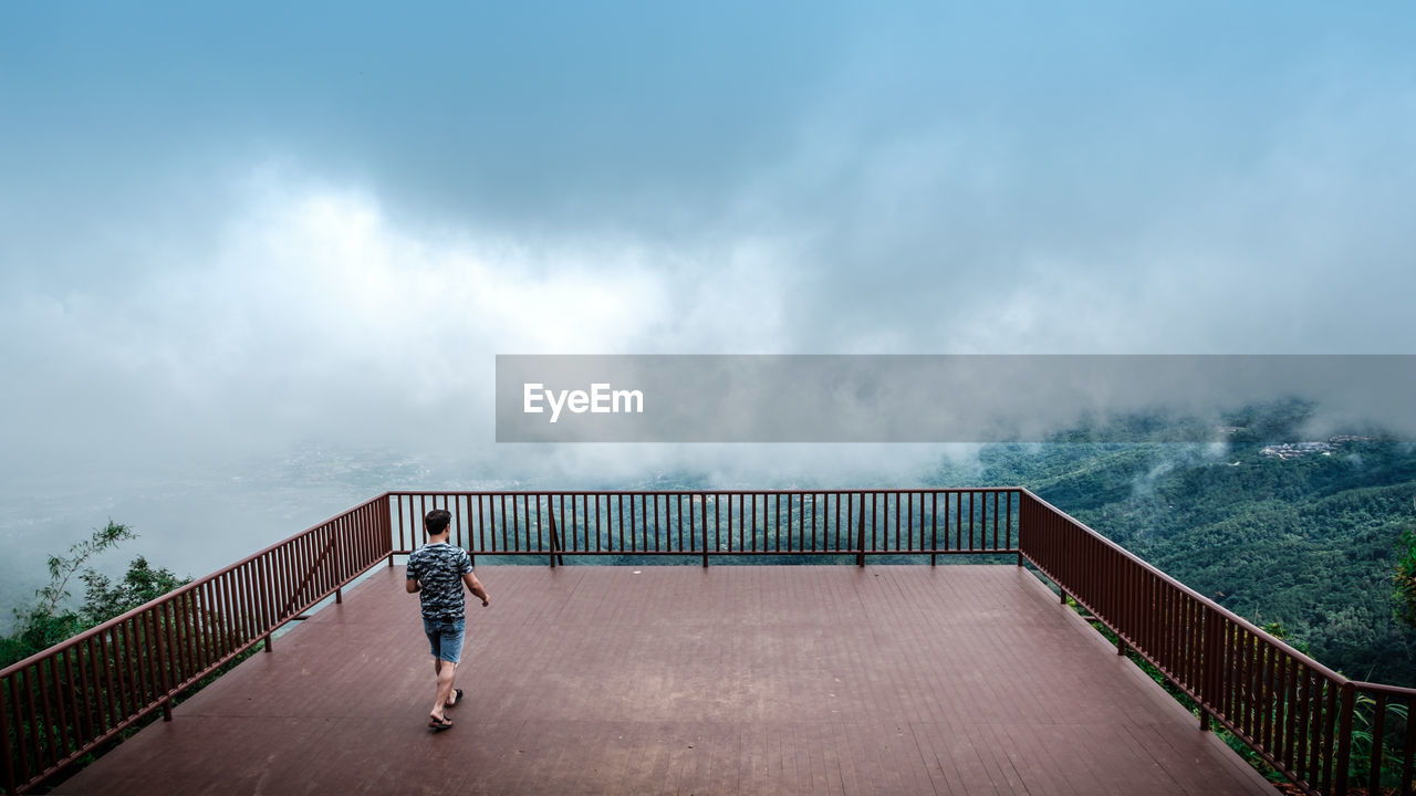 Rear view of man walking at observation point by mountains against cloudy sky