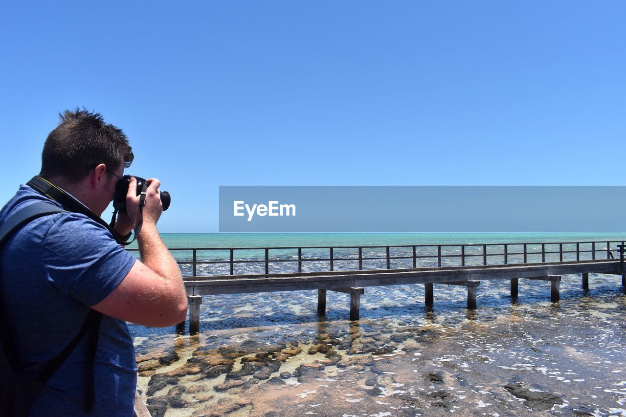 Man photographing at sea against clear blue sky