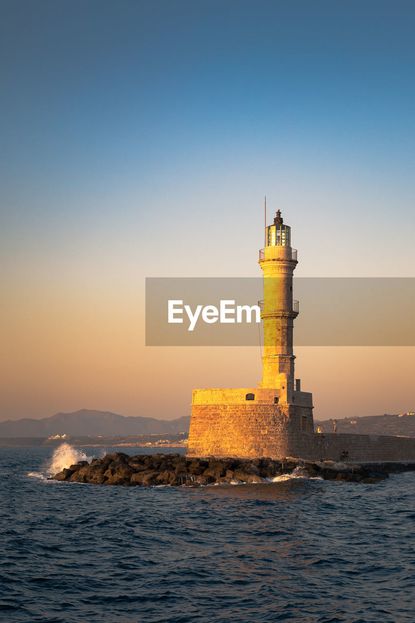 Chania lighthouse view at sunset golden light in the venetian harbour and a wave hitting the cliff 