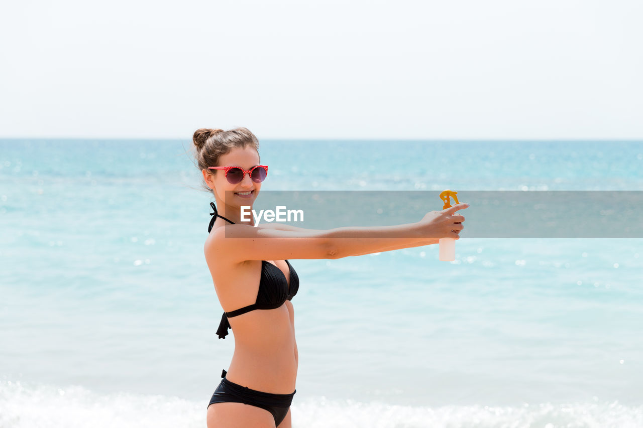 Portrait of smiling bikini woman holding moisturizer bottle while standing at beach