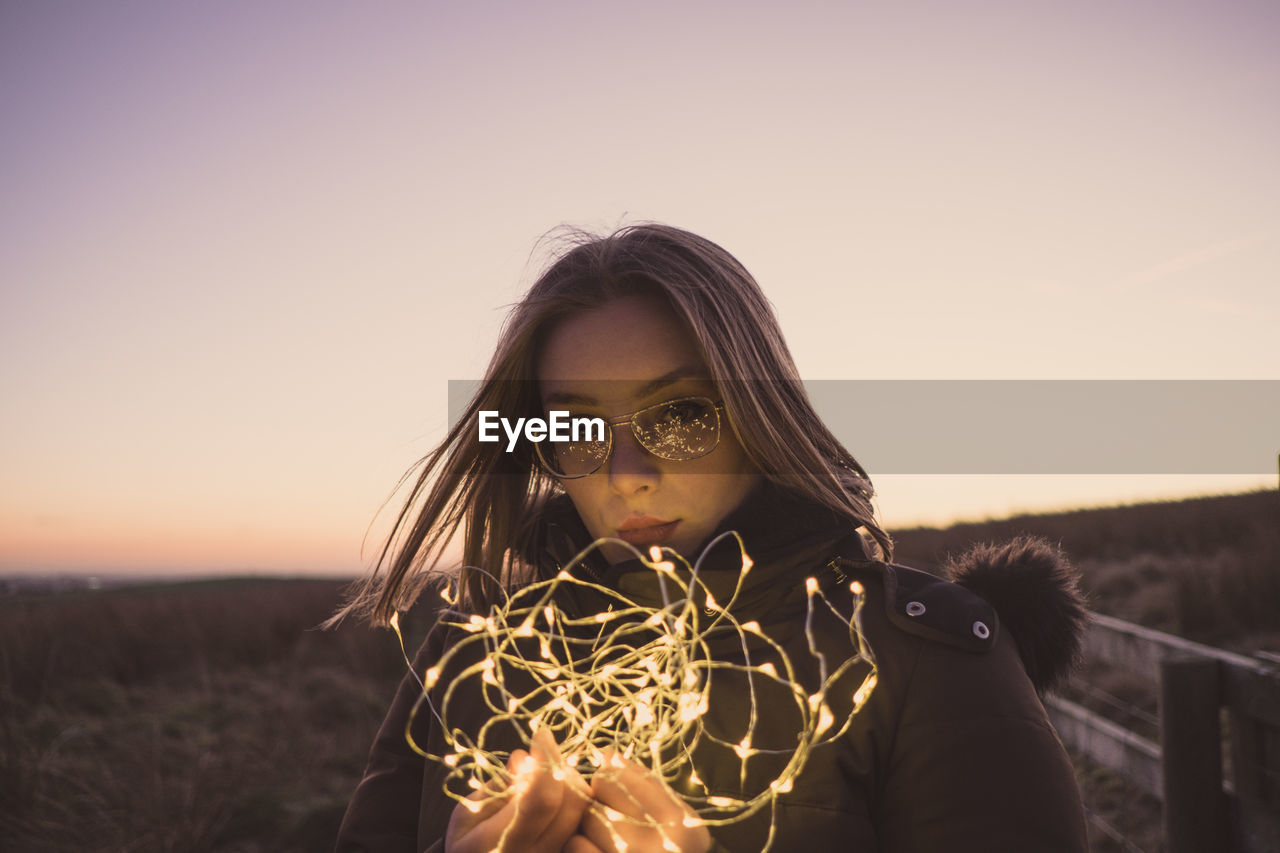 Portrait of woman holding illuminated string lights on field against sky during sunset