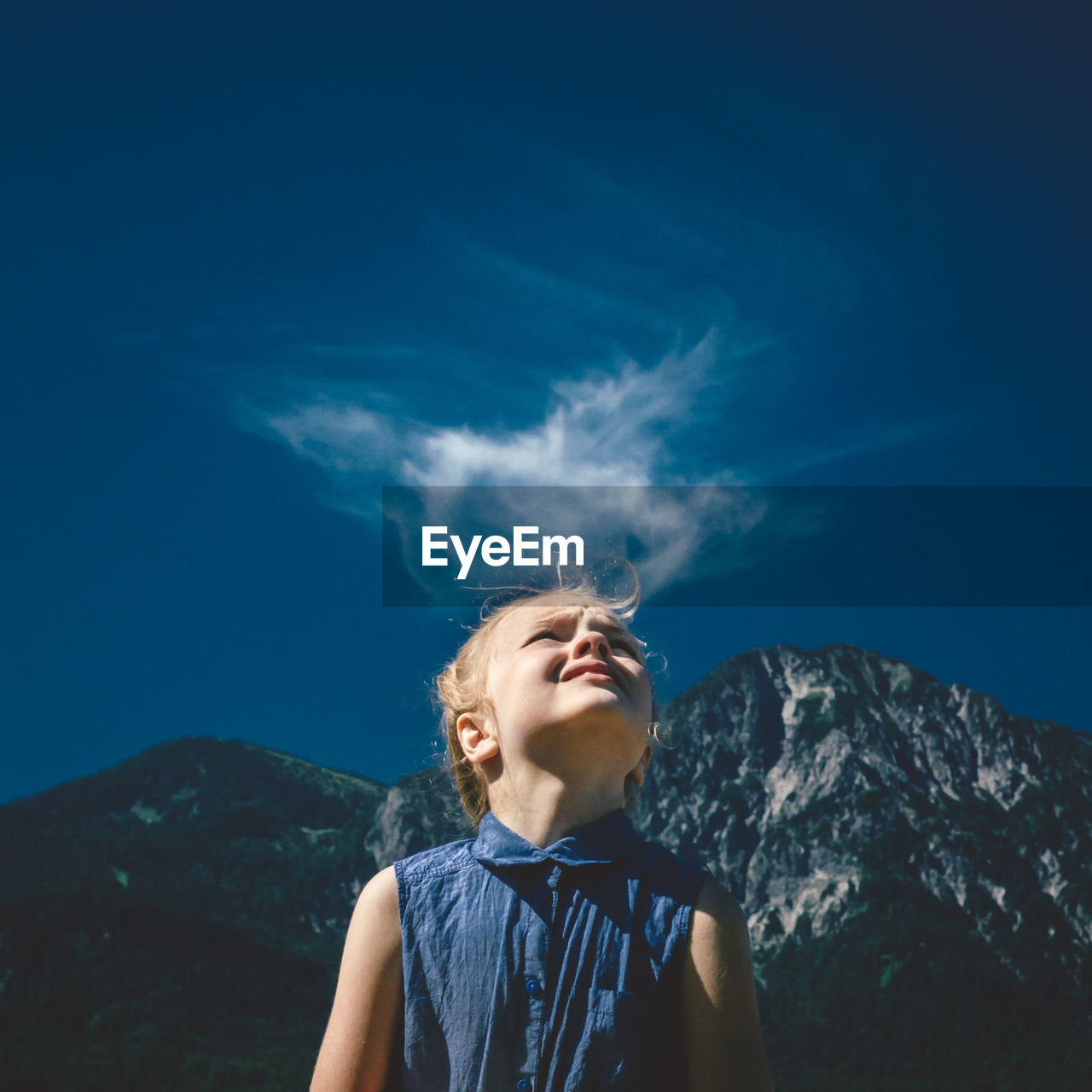 Girl looking up at blue sky with mountain backdrop and cloud above