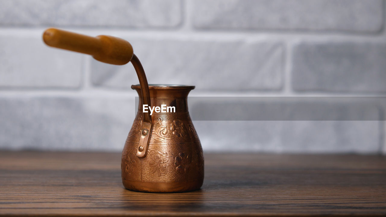 A small copper jezve on a wooden table. in the background is a light stone wall