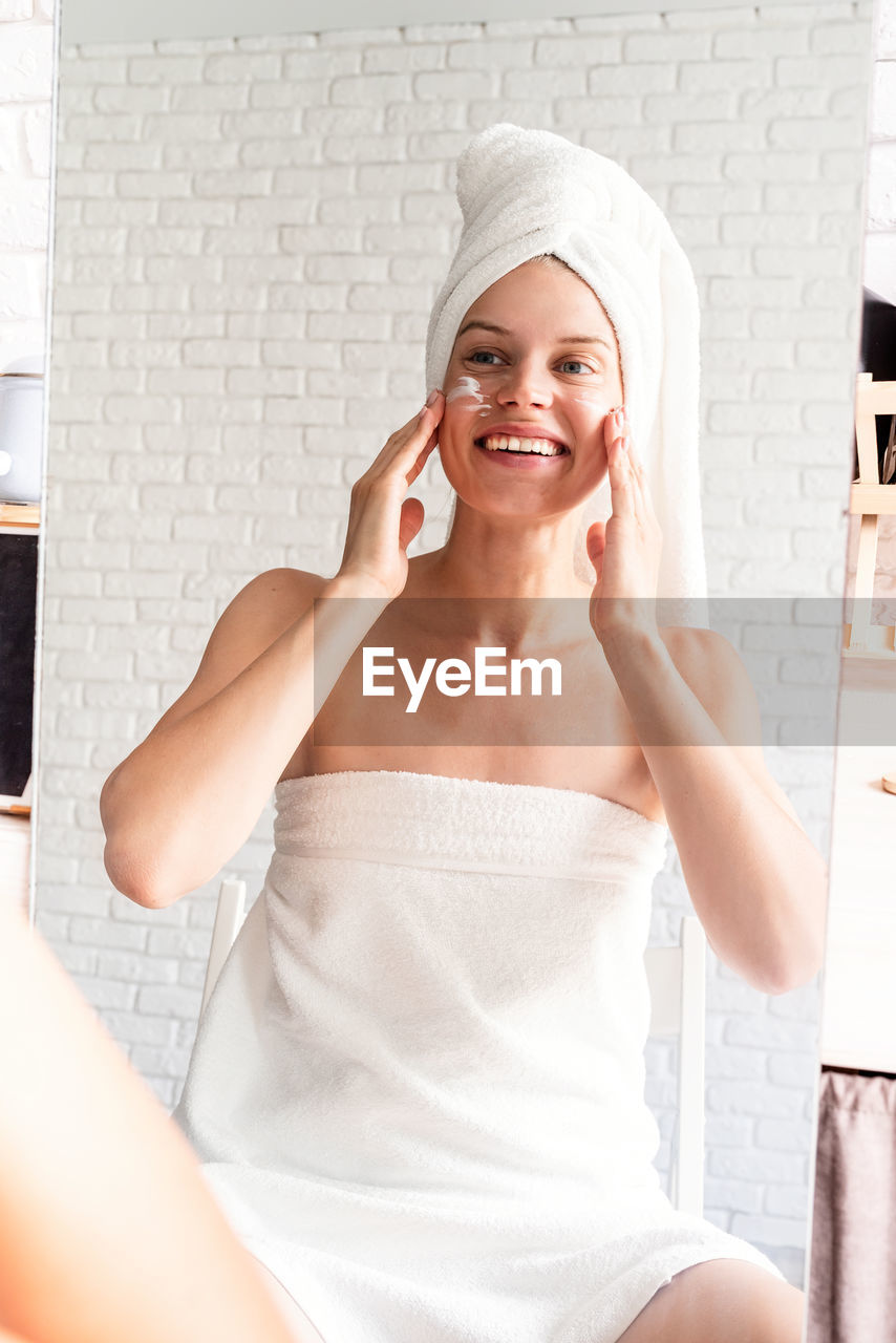 Happy laughing young woman in white bath towels doing morning make up looking at the mirror