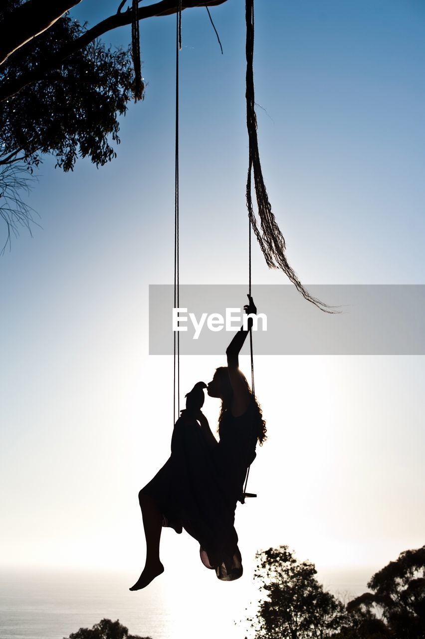 Silhouette of woman kissing bird on swing at sunset