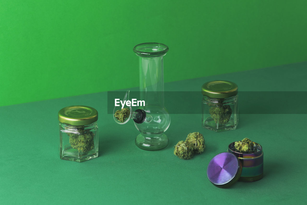 Composition of glass round bong placed near dry cannabis plant in bowl on green surface